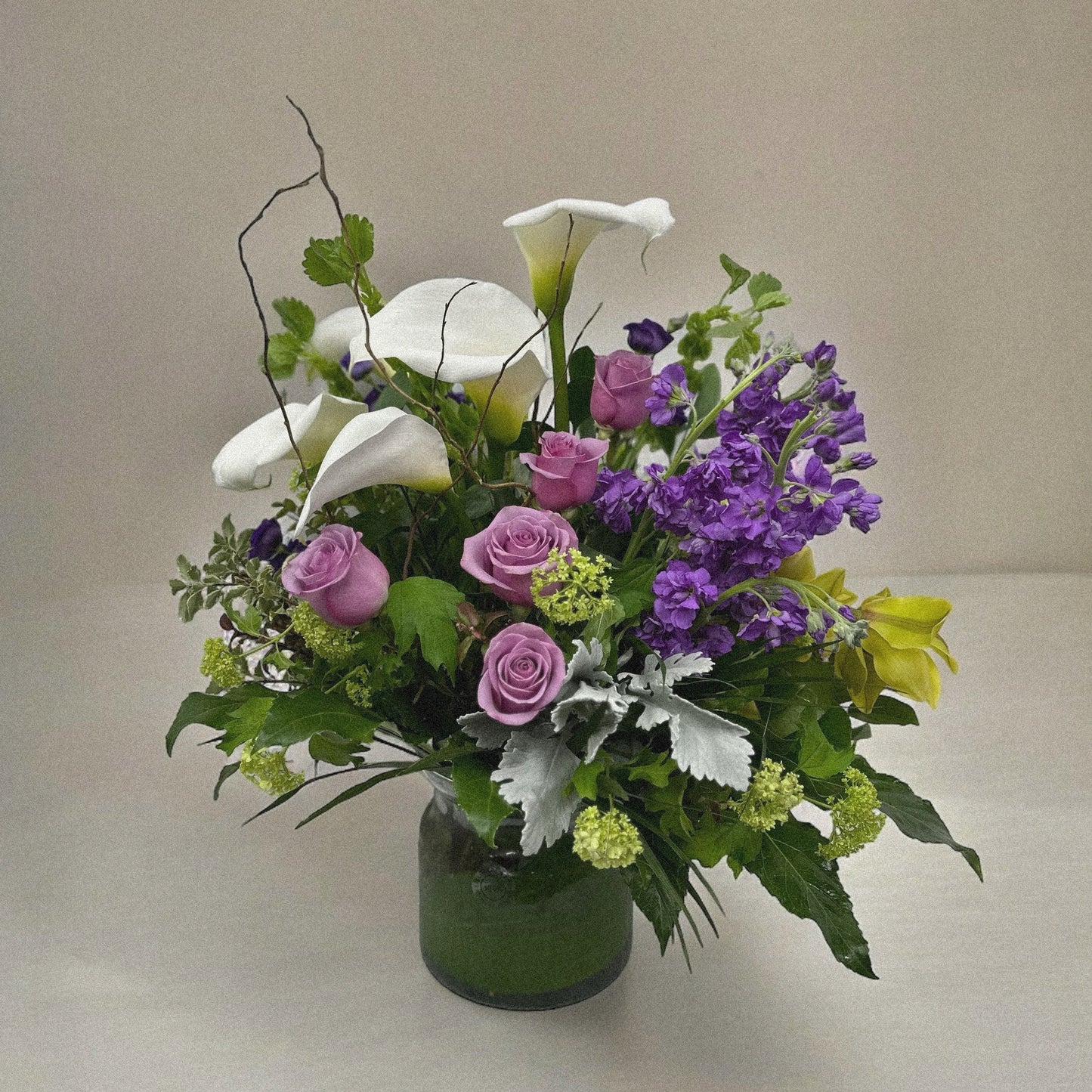 Elegant floral arrangement with white calla lilies, pink roses, purple blooms, and greenery in a clear vase, perfect for all occasions.