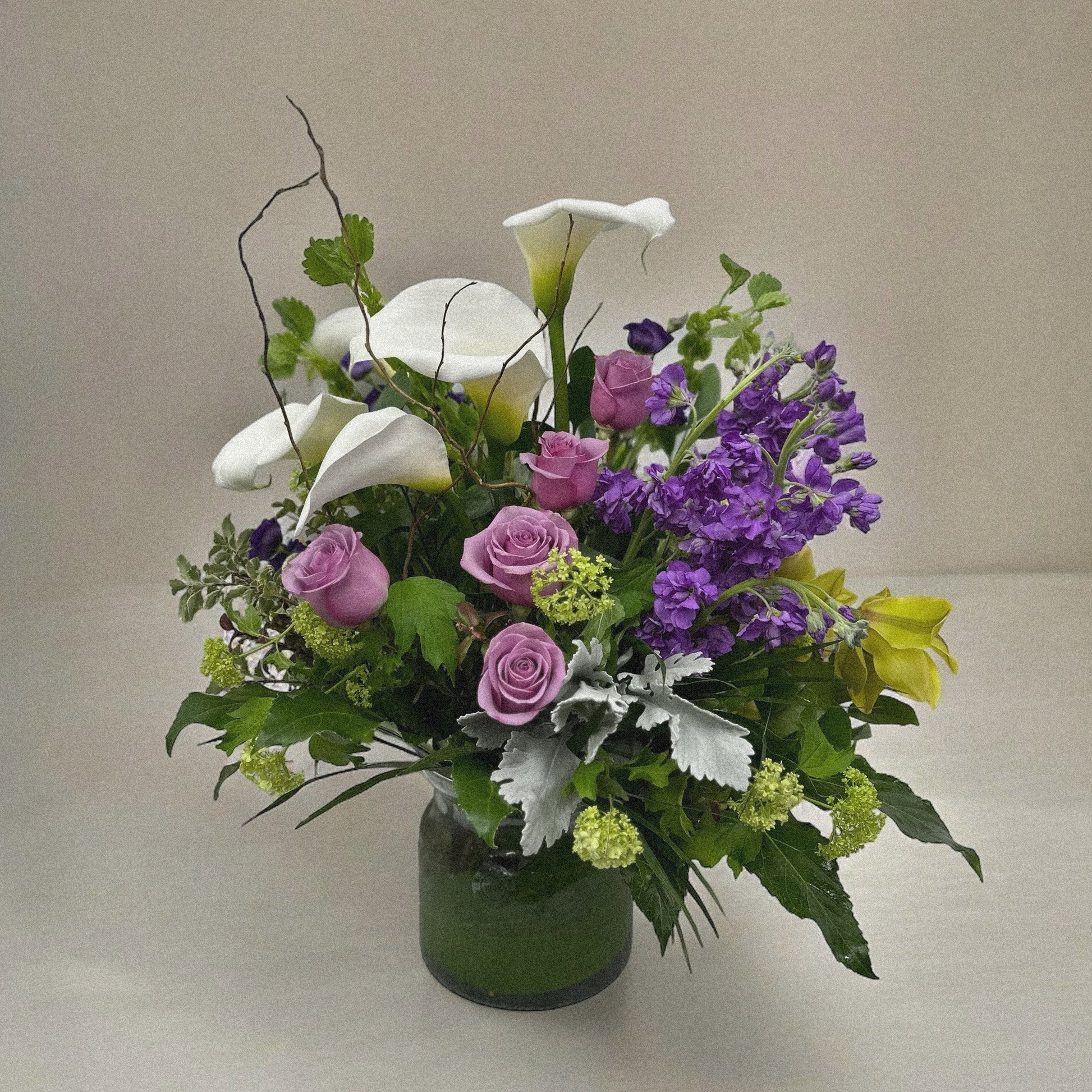 Elegant floral arrangement with white calla lilies, pink roses, purple blooms, and greenery in a clear vase, perfect for all occasions.