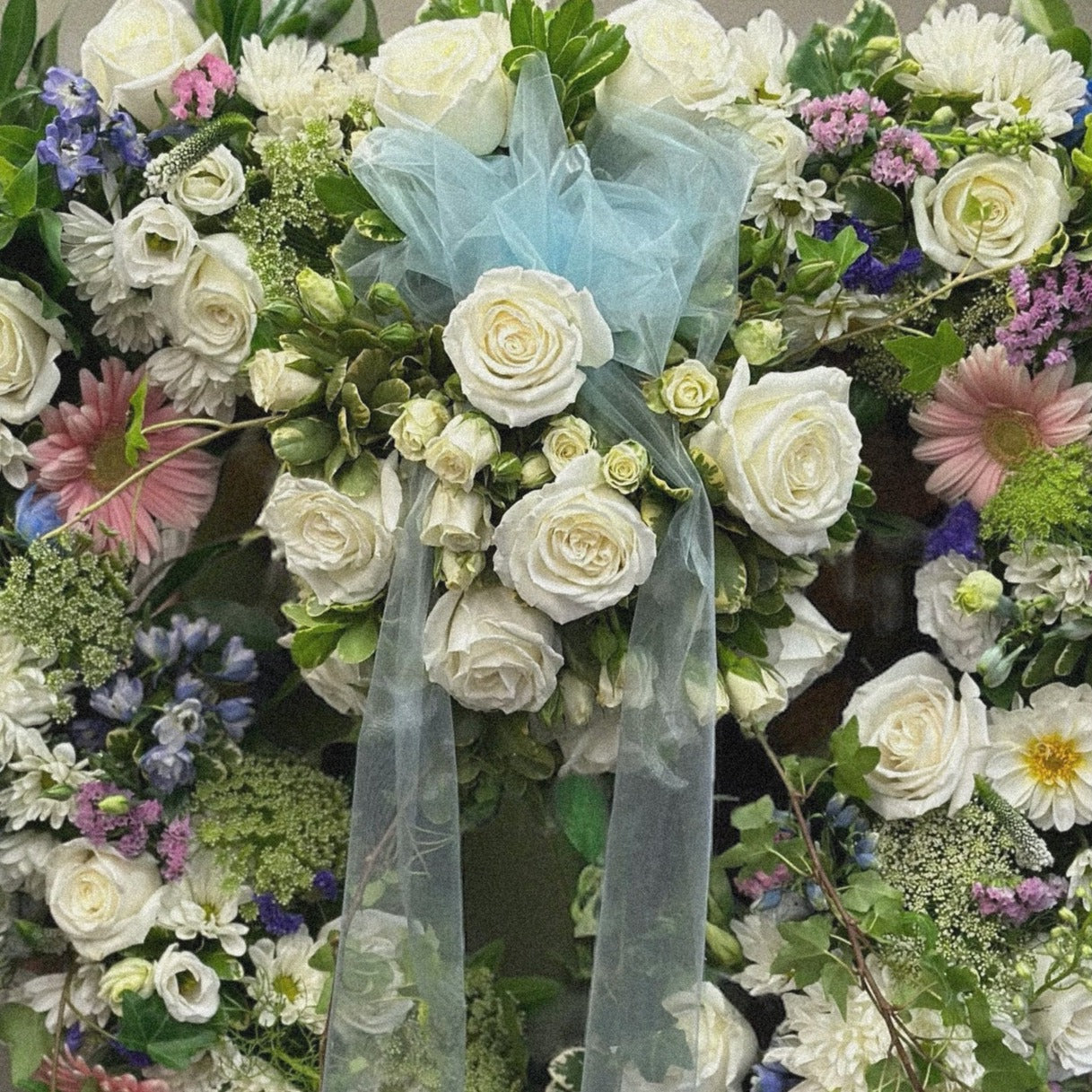 Heartfelt Tribute Wreath with white roses, pink daisies, and blue accents for sympathy and funeral arrangements. Best in Toronto.