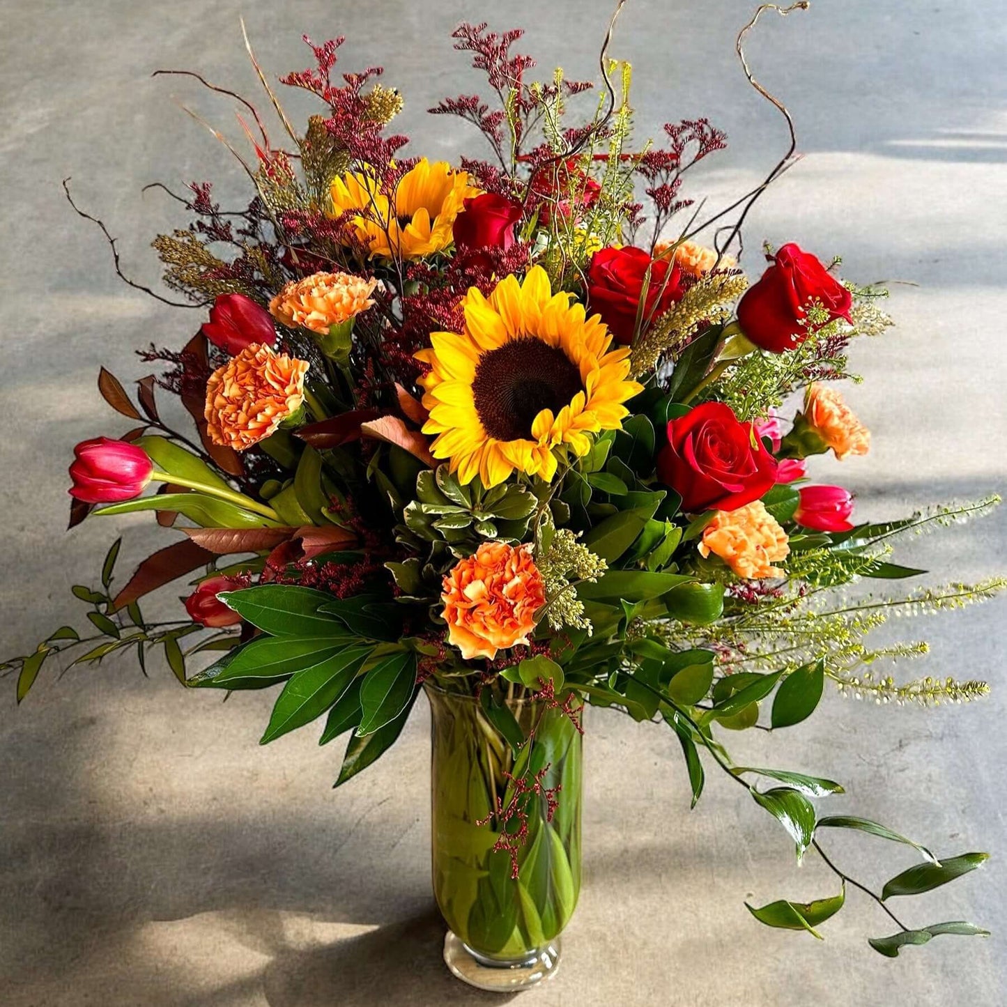 Golden Radiance Bouquet – Sunflower & Rose Fall Arrangement with vibrant sunflowers, red roses, and orange carnations in a glass vase