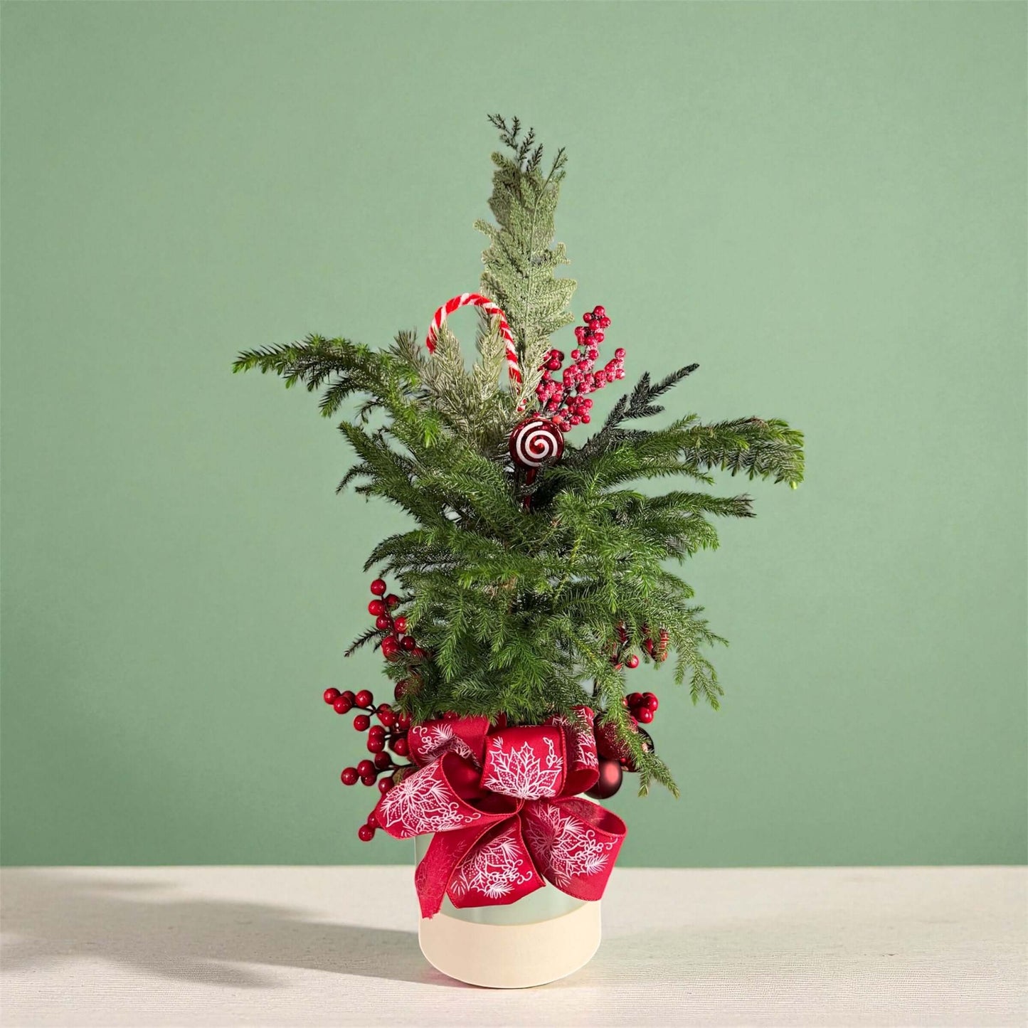 Mini pine tree with red berries, Candy cane ornament, frosted greenery, and red bow in a cream pot.